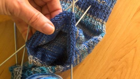 Eine Hand hält eine Socke mit sichtbaren Nadeln, während ein Knäuel blaue Wolle auf einem Tisch liegt.