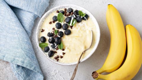 Eine Schüssel Bananen-Joghurt-Eis mit Heidelbeeren, Schokoladenstückchen und Minze, neben frischen Bananen und einem Geschirrtuch.