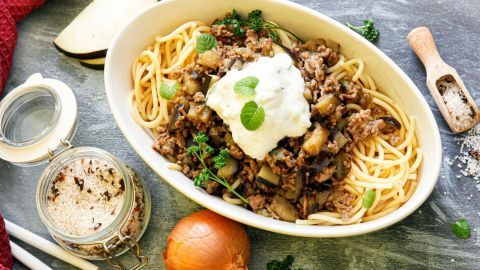 Nudeln mit Hackfleisch und Auberginen sind in einer weißen Schüssel mit Tsatsiki und frischen Kräutern angerichtet.