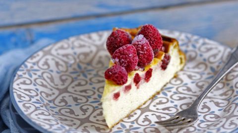Ein Stück Himbeer-Melisse-Käsekuchen liegt auf einem dekorativen Teller und ist mit frischen Himbeeren garniert.