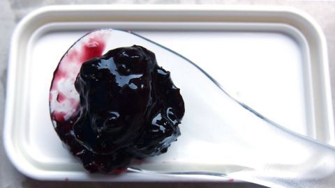 Dunkle Marmelade wird mit einem Löffel aus einer weißen Schale entnommen. Der Hintergrund ist unauffällig.