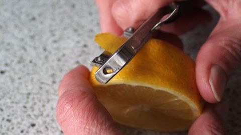 Eine Hand schält sorgfältig eine Zitrone mit einem Messer, um die Schale für Zitronensirup vorzubereiten, auf einer Arbeitsplatte.