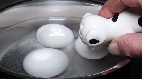 Ein Kochtopf mit drei Eiern in Wasser, eine Hand hält eine Keramikfigur darüber, um beim Eier kochen zu helfen.