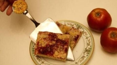 Ein Kuchenstück wird mit einem Tortenheber auf einem Teller serviert, daneben liegen zwei rote Äpfel auf einem Tisch.