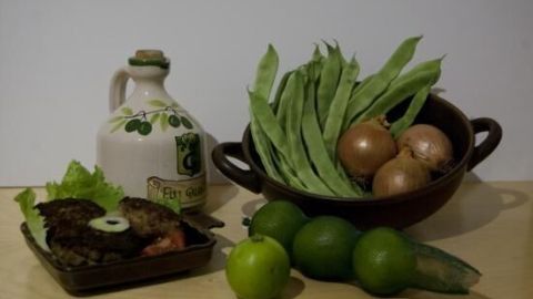 Eine Schüssel mit frischen Bohnen und Zwiebeln steht neben Limetten und einer Flasche Öl auf einem Tisch.