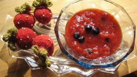Eine Schüssel mit Erdbeermarmelade enthält Aroniabeeren und steht neben crochetierten Erdbeeren auf einem Tisch.