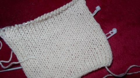 Ein gestricktes, cremefarbenes Rechteck liegt auf rotem Hintergrund, während Gardinenröllchen zur Zählung auf den Nadeln angezeigt werden.