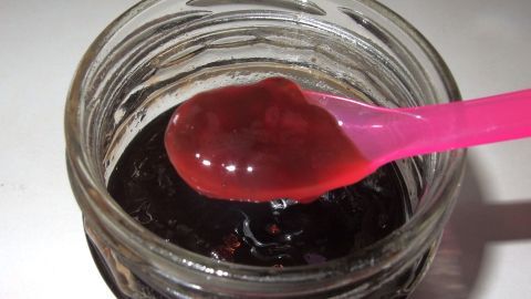 Eine pinke Löffelspitze hebt Marmelade aus einem Glas, die etwas zu dünn wirkt, auf einem hellen Tisch.