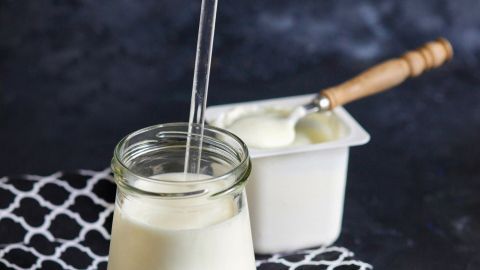 Ein Glas Joghurtmilch steht auf einem Tisch mit einem schwarzen Tuch, daneben ein Becher mit Joghurt und einem Löffel.