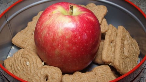 Kleingebäck, Kuchen, Stullen usw. bleiben länger frisch mit einem Apfel. Ein leuchtend roter Apfel liegt in einer Dose, umgeben von knusprigen Keksen, die frisch und appetitlich wirken.