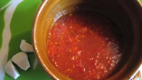 Eine braune Schüssel enthält leuchtend rote Chilisauce; daneben liegen zwei Knoblauchzehen auf einem grünen Untergrund.