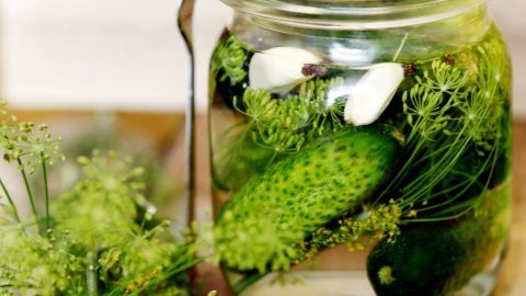 Ein Glas mit Gewürzgurken und Dill ist im Vordergrund, während eine Gurke ins Glas eingetaucht wird, um extra pikant eingelegt zu werden.