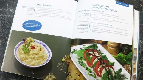 Halte dein Kochbuch sauber, indem du deine Lieblingsrezepte vor Flecken schützt. Verwende als Schutz einen schlichten Untersetzer, während du dich an der Zubereitung von Baba Ganoush oder Caprese und Mango-Mozzarella inspirieren lässt. Ein Kochbuch liegt offen auf einem Marmortisch. Links zeigt es ein Bild von Baba Ganoush, rechts Caprese und Mango-Mozzarella.