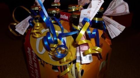 Ein Sechserpack Bierflaschen, dekoriert mit bunten Bändern und 10-Euro-Geldscheinen, im Halbkarton auf Teppichboden.