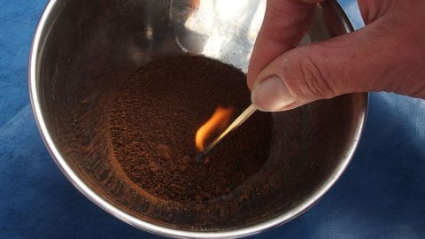 In einer Schüssel mit Kaffeepulver zündet eine Hand mit einem Streichholz das Pulver an. Ein experimenteller Kontext.