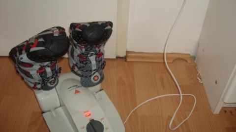 Ein Schuhtrockner trocknet nasse Schuhe auf Holzboden. Er ist eingesteckt und zeigt eine leuchtende Kontrollleuchte.