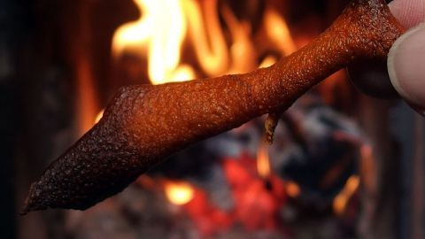 Ein Natur-Feueranzünder wird in der Hand gehalten, während im Hintergrund ein offenes Feuer brennt und Glut leuchtet.