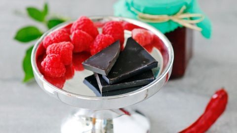 Eine silberne Schale präsentiert frische Himbeeren und Schokolade, daneben ein Glas mit Himbeermarmelade auf einem hellen Hintergrund.