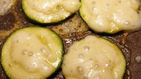 Runde Zucchini-Scheiben brutzeln in einer Pfanne, bedeckt mit geschmolzenem Käse, in einer herzhaften Küchenumgebung.
