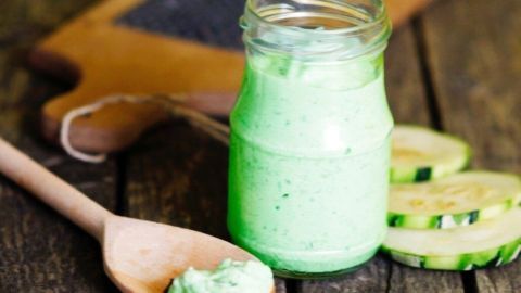 Ein Glas mit grünem Zucchini-Pesto steht auf einem rustikalen Holztisch, daneben liegt ein Löffel mit etwas Pesto und Gurkenscheiben.