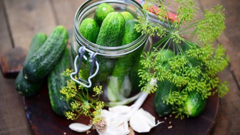 Eingemachte saure Gurken in einem Glas, umgeben von frischen Gurken, Dill und Knoblauch auf einem Holzbrett.