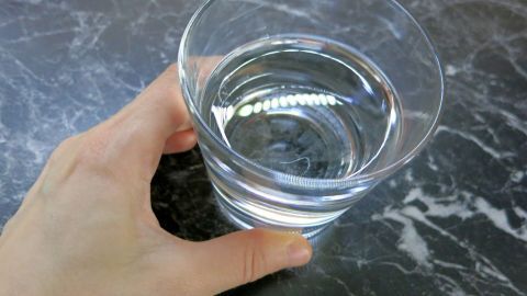Ein einfacher Trick gegen Schluckauf: Trink ein Glas Wasser auf dem Kopf stehend! Stabilisiere ein Glas Wasser auf einem Tisch und beug dich nach unten, um langsam von der anderen Seite des Glases zu trinken. Eine Hand hält ein Glas mit Wasser auf einem marmorgefärbten Tisch. Dieses könnte im Kontext Schluckauf besiegen helfen.