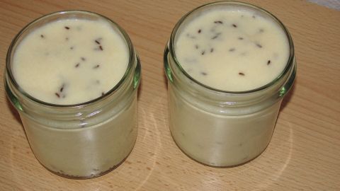 Zwei Gläser mit Kochkäse stehen auf einem Holzbrett. Die cremige Masse hat eine leicht gesprenkelte Oberfläche.