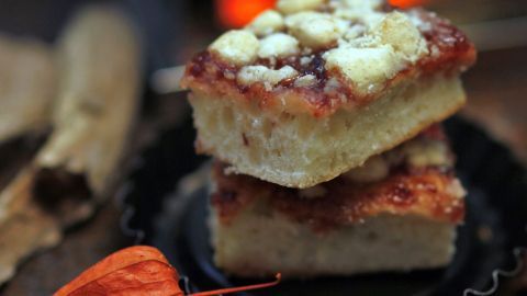 Zwei Stücke Zwetschgenkuchen mit Streuseln liegen übereinander auf einer dunklen Unterlage, umgeben von orangefarbenen Dekorationen.