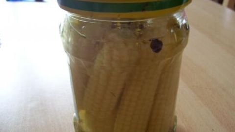 Ein Glas mit eingelegten kleinen Maiskölbchen steht auf einem Tisch, umgeben von einer hellen, gemütlichen Küchenszene.