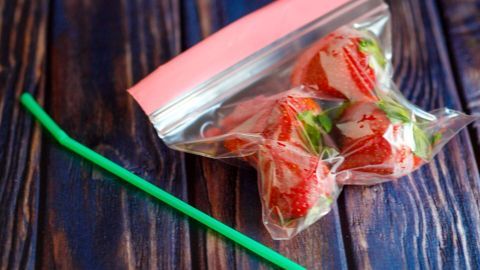 Lebensmittel in einem Zip-Beutel mithilfe eines Strohhalms vakuumieren. Eine Tüte mit frischen Erdbeeren liegt auf einem Tisch, während ein grüner Strohhalm daneben für das Vakuumieren verwendet wird.