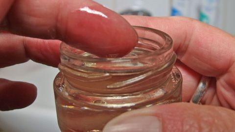 Eine Hand greift nach Olivenöl in einem kleinen Glasbehälter, während eine Badezimmerumgebung sichtbar ist.