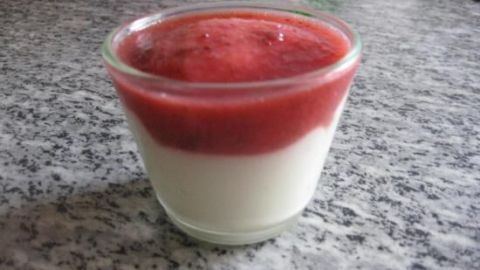 Ein Dessert mit weißer Schokolade und einer fruchtigen Erdbeersoße in einem Glas auf einer grauen Arbeitsplatte.