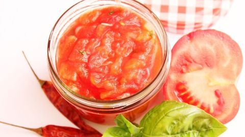 Ein Glas Tomatensoße steht auf einem Tisch, umgeben von frischen Tomaten, Basilikum und Chilischoten.