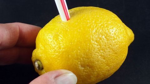 Eine Hand hält eine Zitrone, in die ein Strohhalm gesteckt ist, während die Zitrone auf einem dunklen Untergrund liegt.