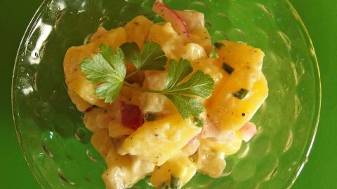 Ein bunter Kartoffelsalat wird in einer durchsichtigen Schüssel serviert, umgeben von frischem Gemüse und Kräutern.