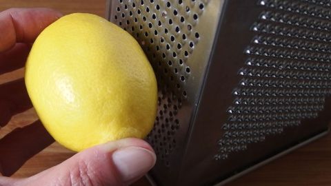 Eine Hand hält eine frische Zitrone, während sie sie über eine Reibe in der Küche reibt, um Zitronenschale zu gewinnen.