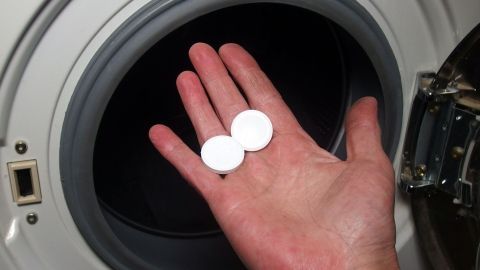 Eine Hand hält zwei weiße Tabletten über einer Waschmaschine, um Sportwäsche frisch zu halten und Gerüche zu vermeiden.