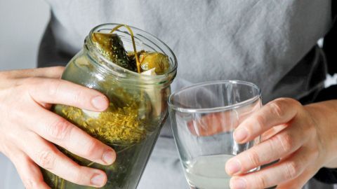 Ein Glas wird mit Gurkenwasser gefüllt, während ein Glas mit klarem Wasser in der anderen Hand gehalten wird.