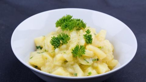 Ein Kartoffelsalat in einer weißen Schüssel, garniert mit frischer Petersilie, steht vor einem dunklen Hintergrund.