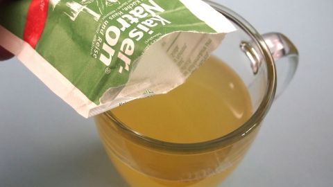 Eine Hand schüttet ein Päckchen Pulver in ein Glas mit goldenem, spritzigem Eistee, vorbereitet zur Erfrischung.