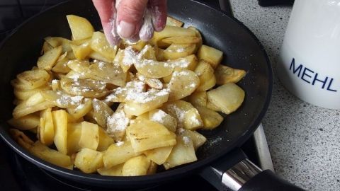 Um krosse Bratkartoffeln zu bekommen, die Kartoffeln immer wieder mit etwas Mehl bestäuben. In einer Pfanne braten krossen Kartoffeln, während eine Hand Mehl hinzufügt, um die knusprige Textur zu verbessern.