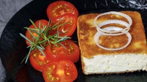 Gebackener Schafskäse liegt auf einem schwarzen Teller, umgeben von frischen Tomatenscheiben und Zwiebelringe.