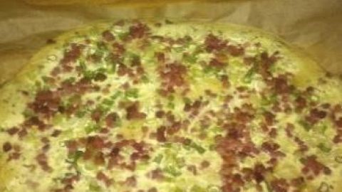 Ein runder Flammkuchen mit Hefeteig, belegt mit Speckwürfeln und Frühlingszwiebeln, ruht auf Backpapier auf Holzoberfläche.