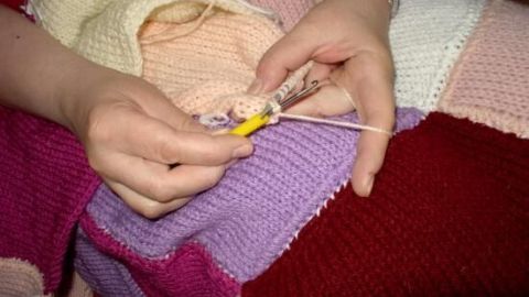 Gestrickte Hände nutzen eine Häkelnadel, um an einem bunten, gestickten Quilt weiterzuarbeiten.