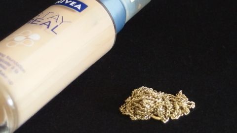 Eine goldene Kette liegt auf einem schwarzen Untergrund, während daneben eine Flasche Make-up aufrecht steht.