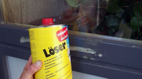 Ein Hand hält eine gelbe Dose mit "Löser" neben einem Fensterrahmen, der Klebereste aufweist, in einem hellen Innenraum.