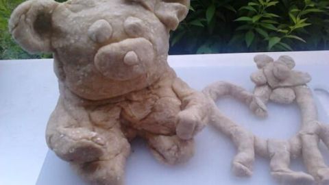 Ein sitzender, kneteförmiger Bär mit großen Ohren hält eine kleine Blume in der Hand, umgeben von grünem Pflanzenhintergrund.
