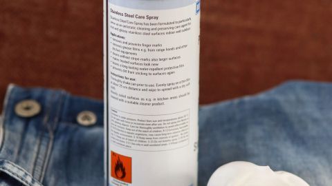 Eine Spraydose mit Backofenschaum liegt auf einer Jeans, während der Schaum bereit ist, um Farbflecken zu entfernen.