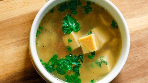 Eine helle Suppe mit grüner Petersilie und Käsewürfeln wird in einer weißen Schüssel auf einem Holzbrett serviert.