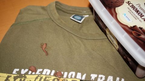Ein olivgrünes T-Shirt mit einem braunen Schokoladeneisfleck liegt neben einer Eisverpackung auf einem Tisch.